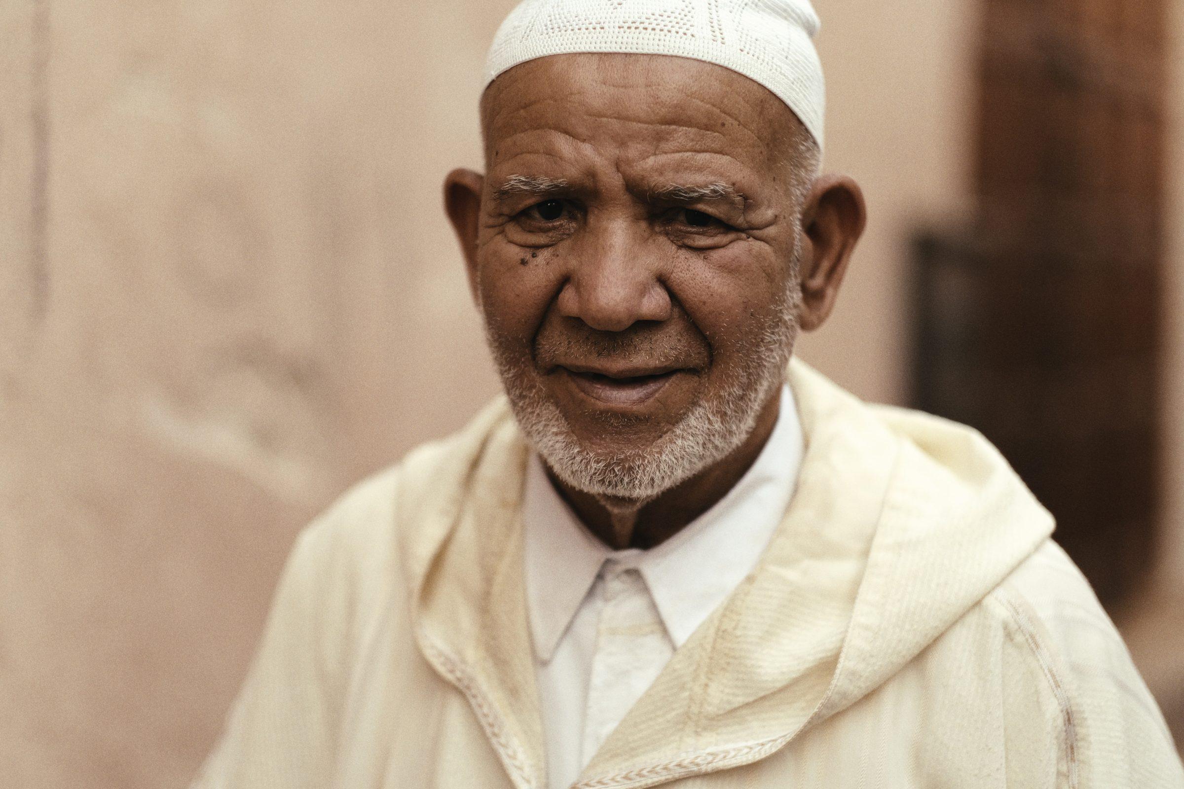 Close portrait of an older man in a white cap and robe, grey beard, a soft wall behind him