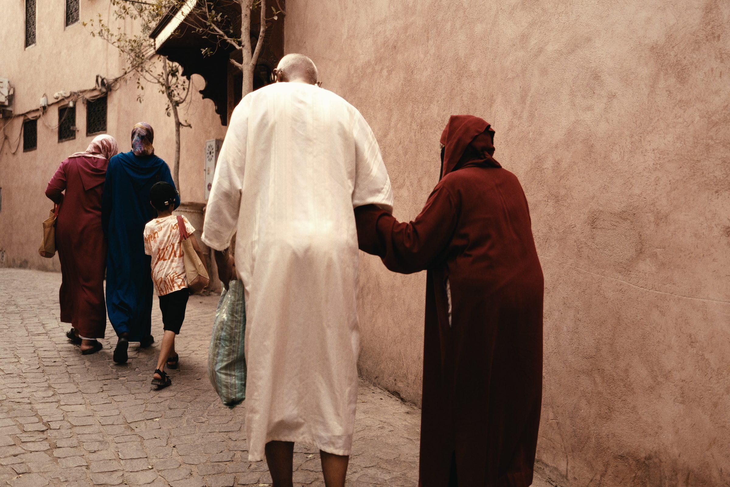 Two people in long robes walking along a terracotta wall, one steadying the other by the arm, a family ahead of them