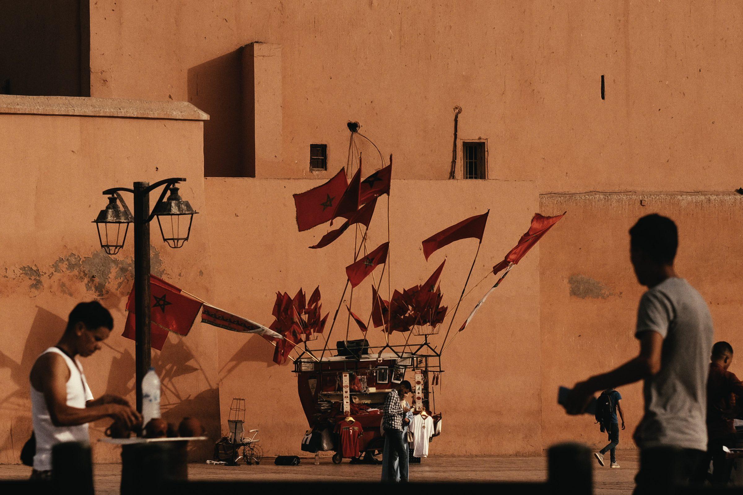 Red flags on a small stall against an ochre wall, figures passing in silhouette