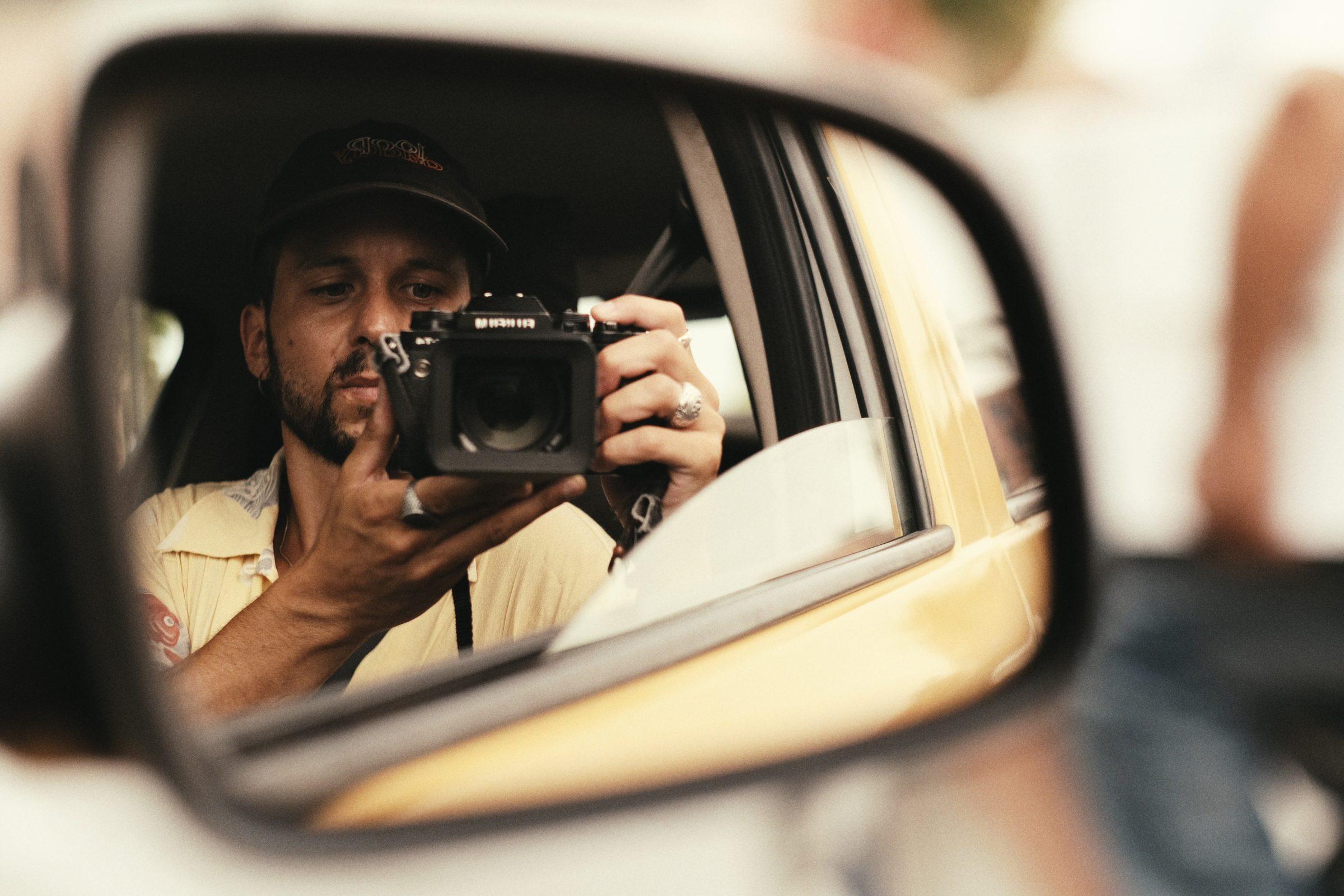 A man with a camera reflected in a car's wing mirror, a yellow taxi beside him