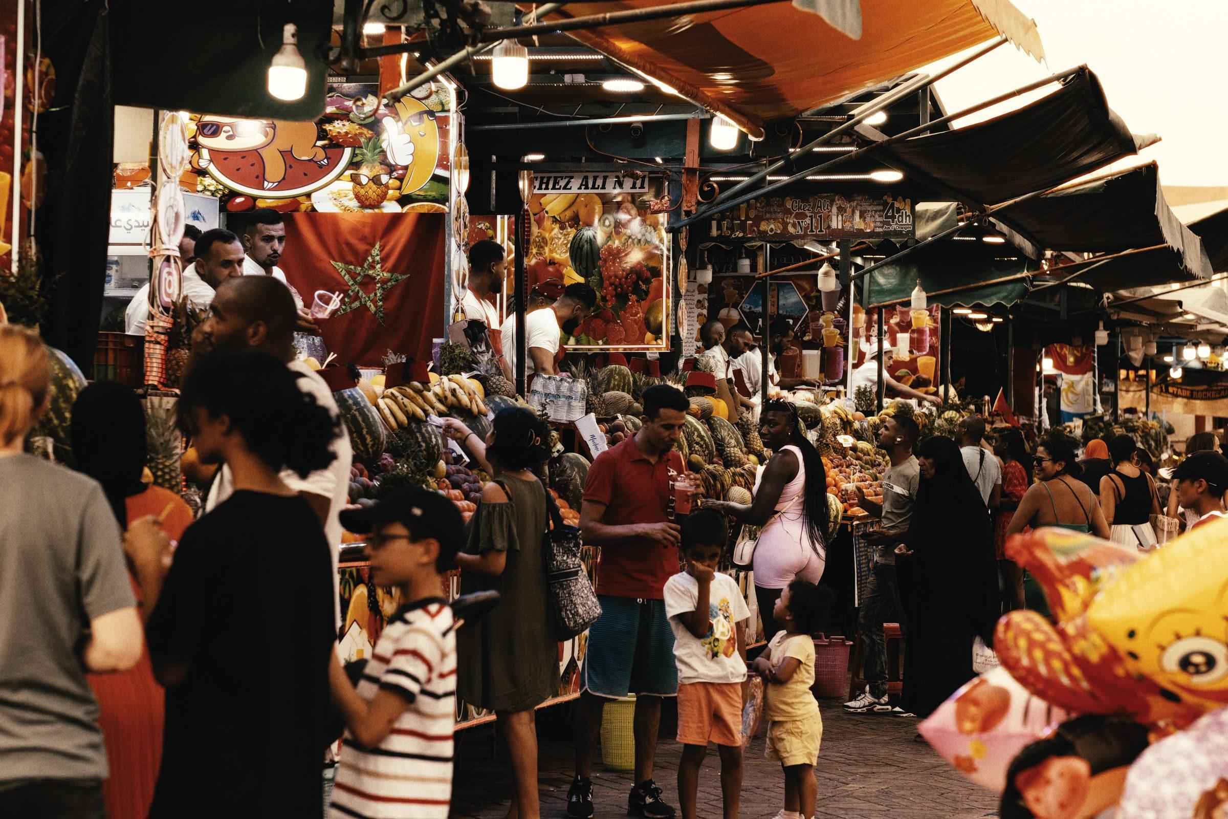 A crowded market lane under strings of lights, children and shoppers among the stalls