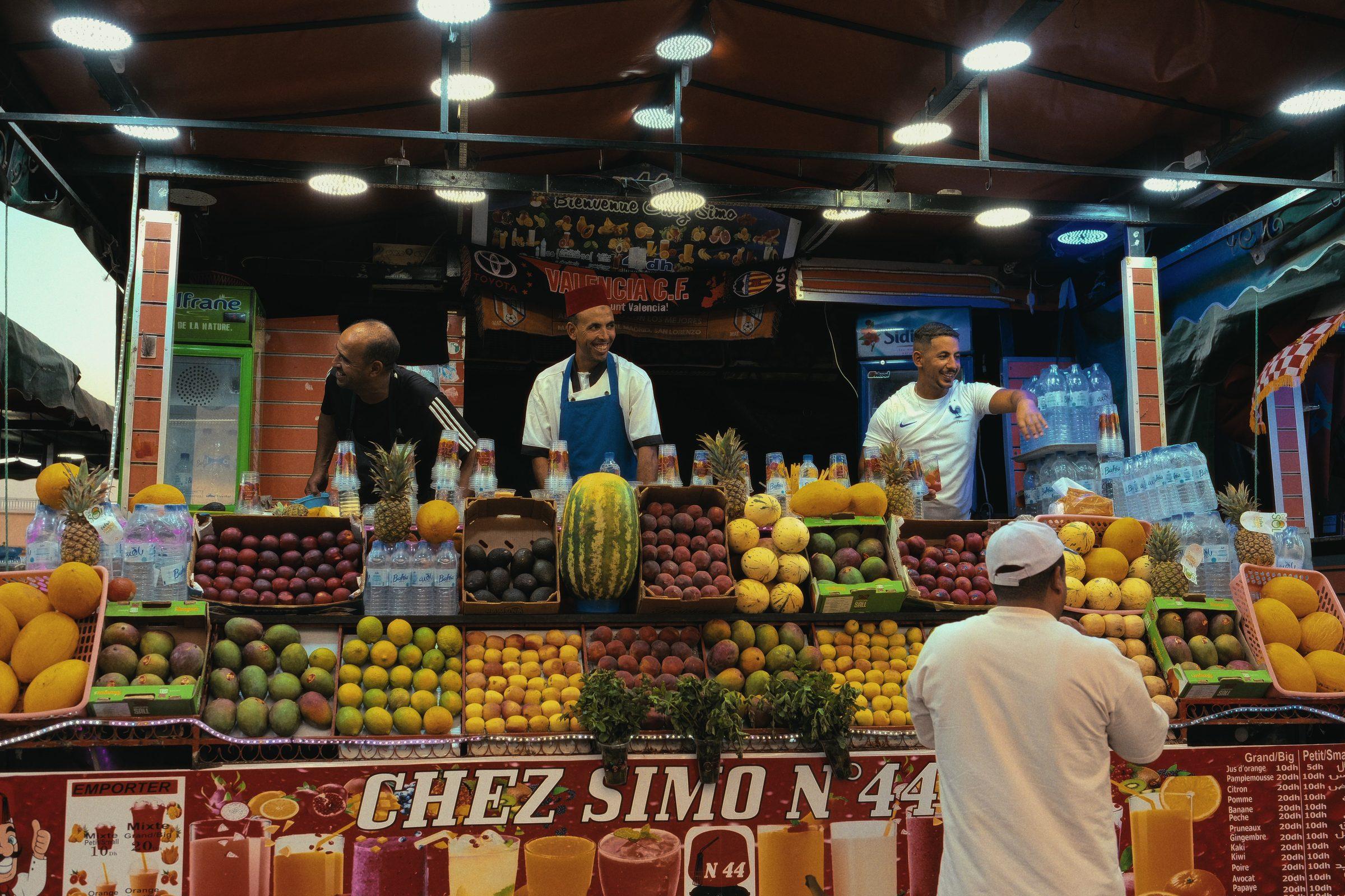 A juice stall stacked with fruit, three men behind the counter and a customer in white in front