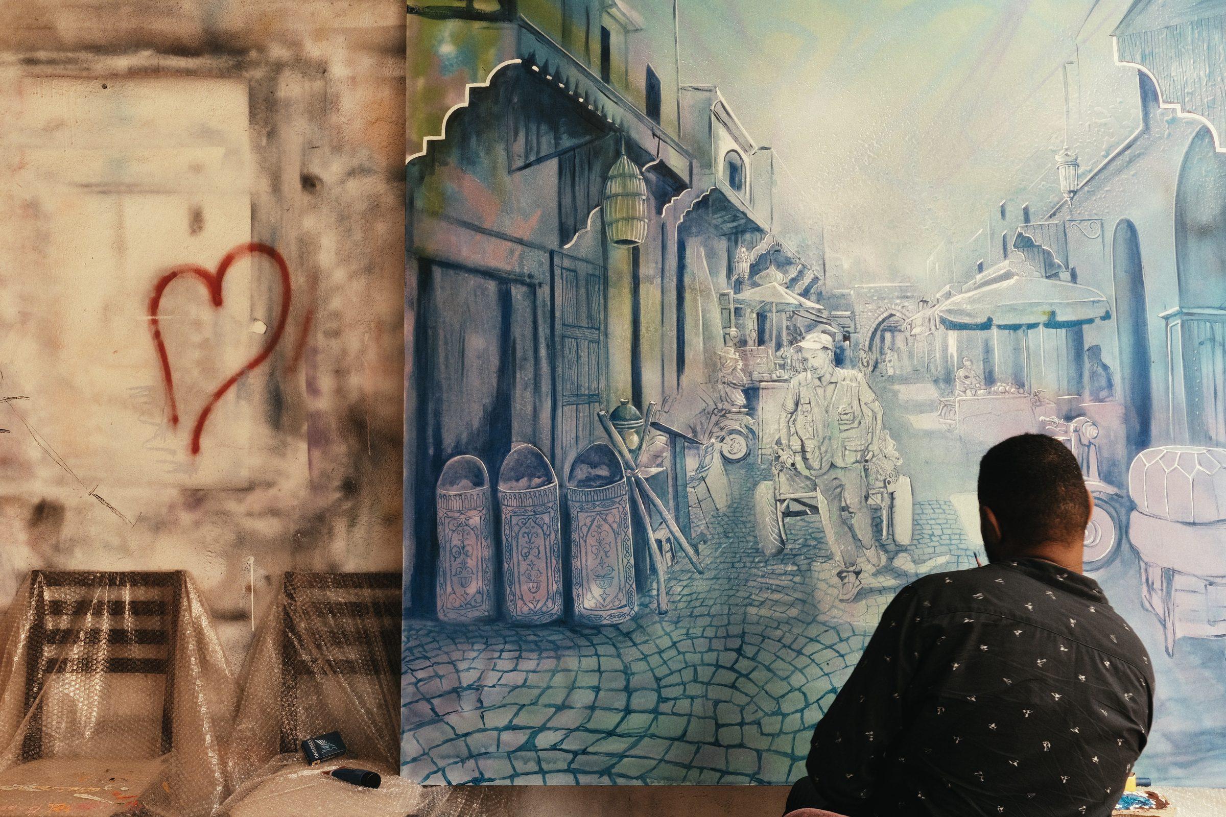 A man seated with his back turned, looking at a painted mural of an old street beside a red heart on the wall