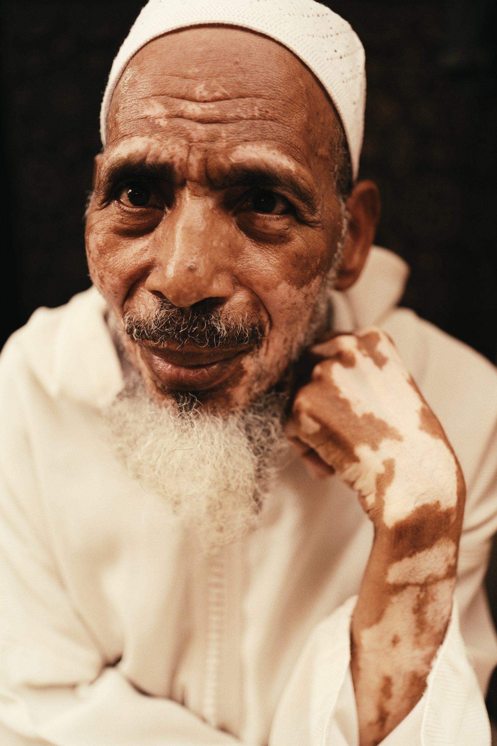 Portrait of an older man with a white beard, in a white robe, resting his chin on his hand