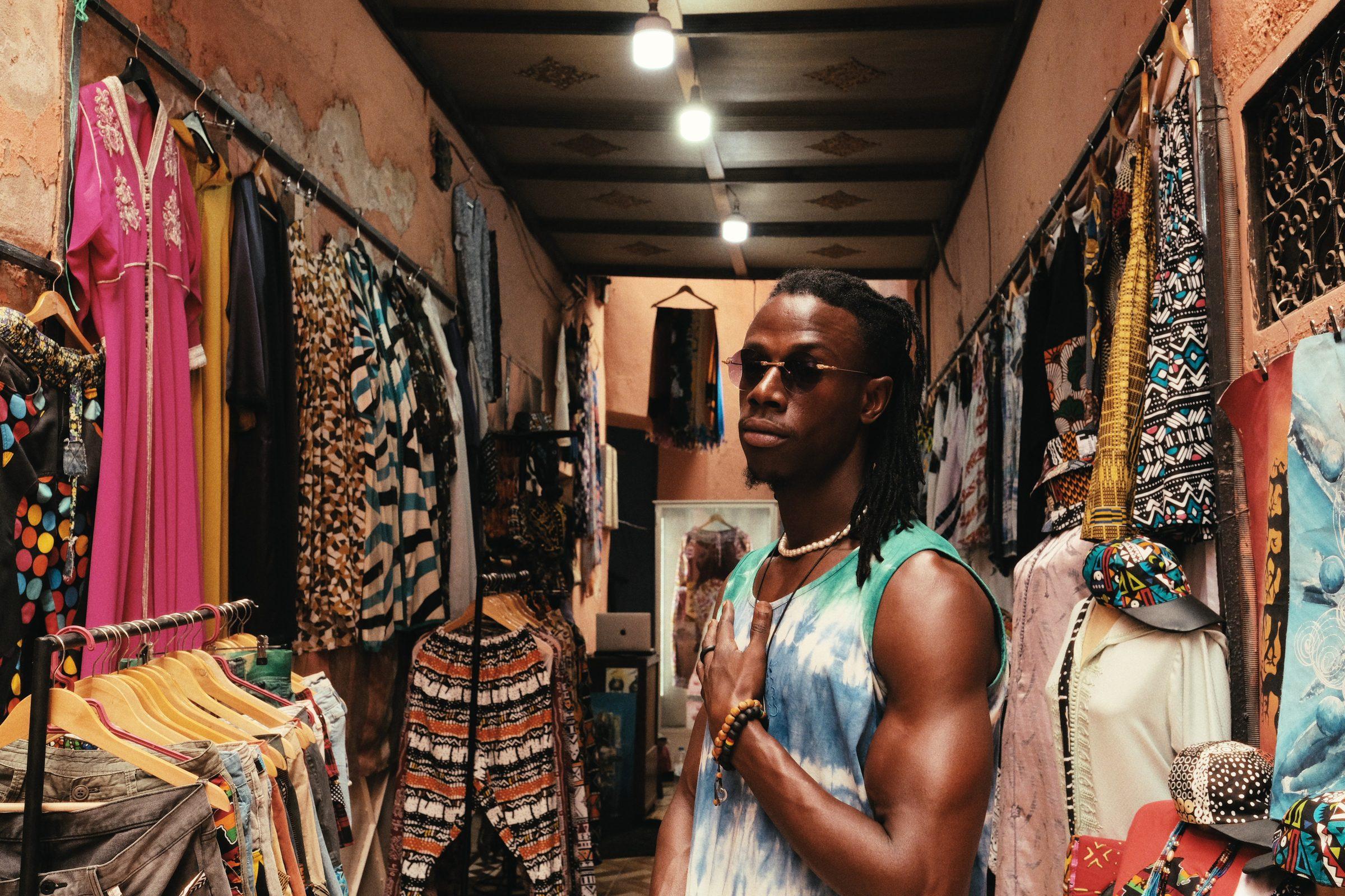 A man in a tie-dyed vest standing in a clothing shop hung with kaftans and patterned trousers