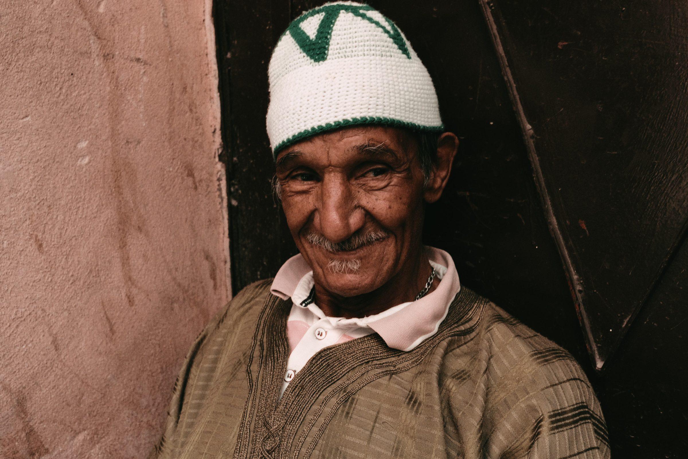 A smiling man in a green and white knitted cap, leaning out of a shadowed doorway against a pink wall