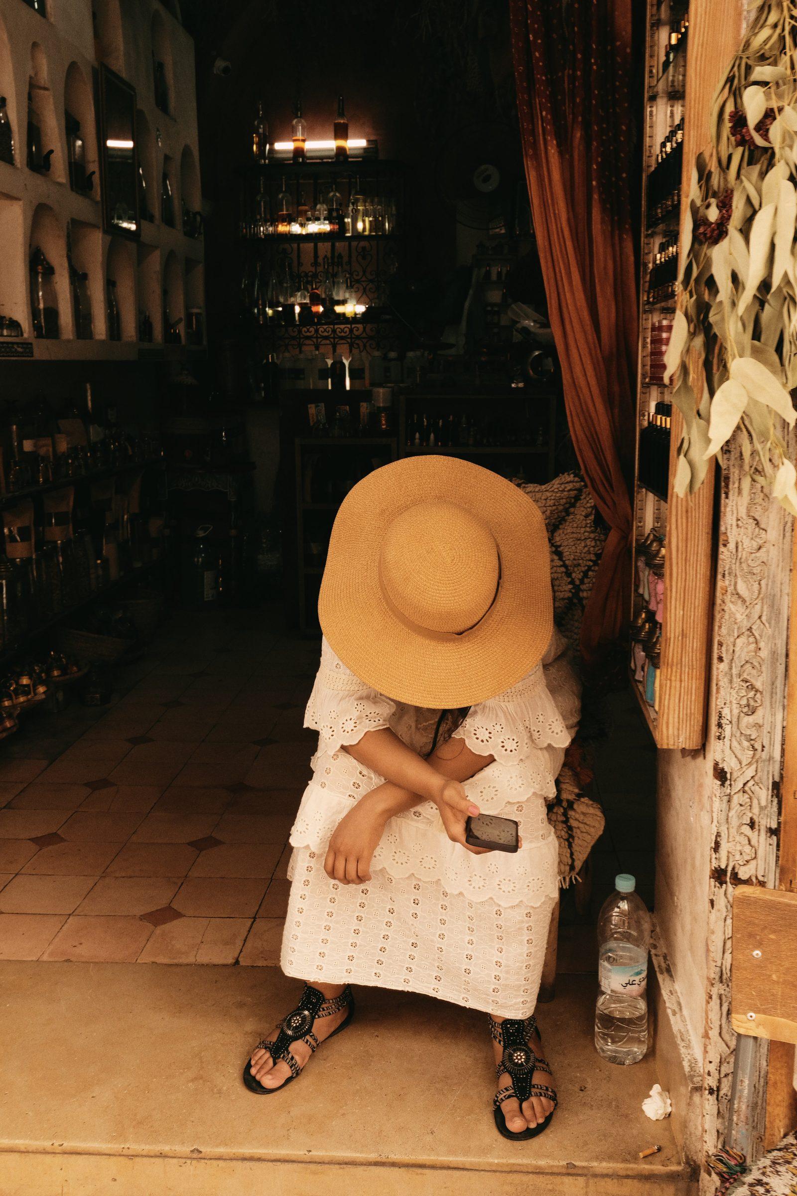 A woman in a sun hat and white dress sitting in a shop doorway, her face hidden by the hat's brim