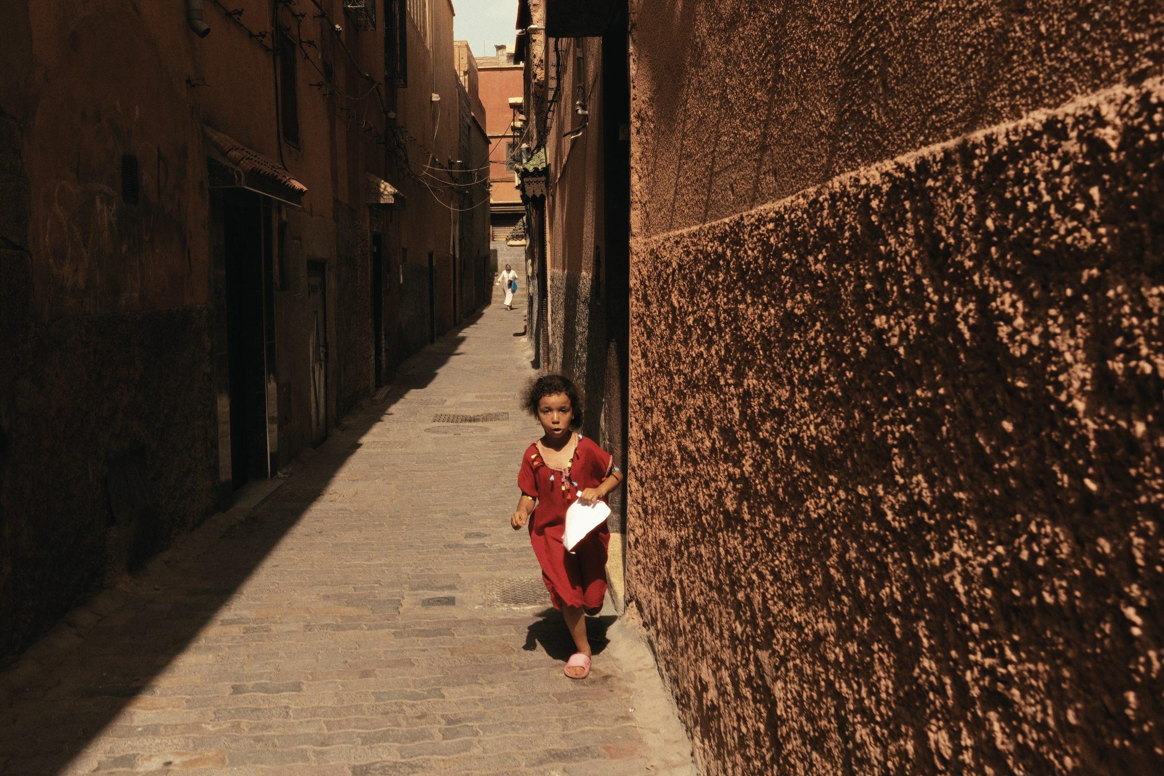 A small girl in a red dress running down a narrow, sunlit lane