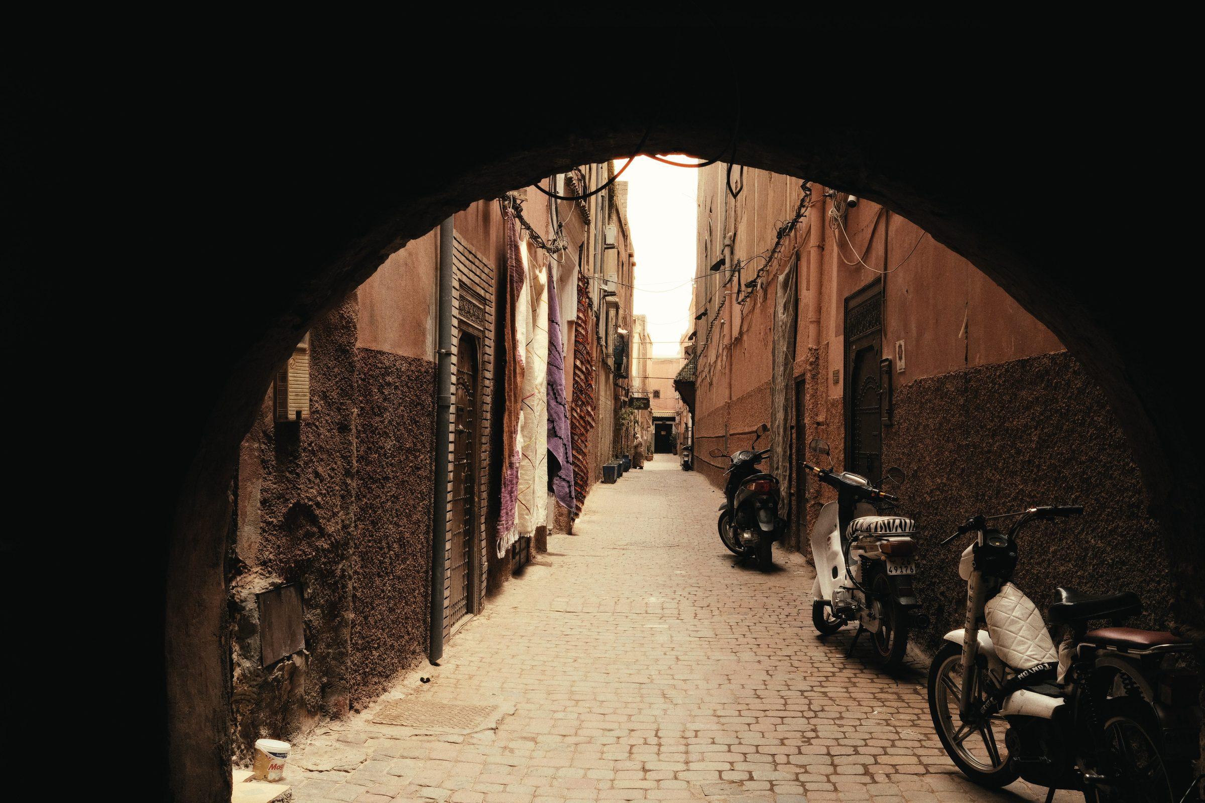 A sunlit alley seen through a dark stone archway, scooters parked along the wall