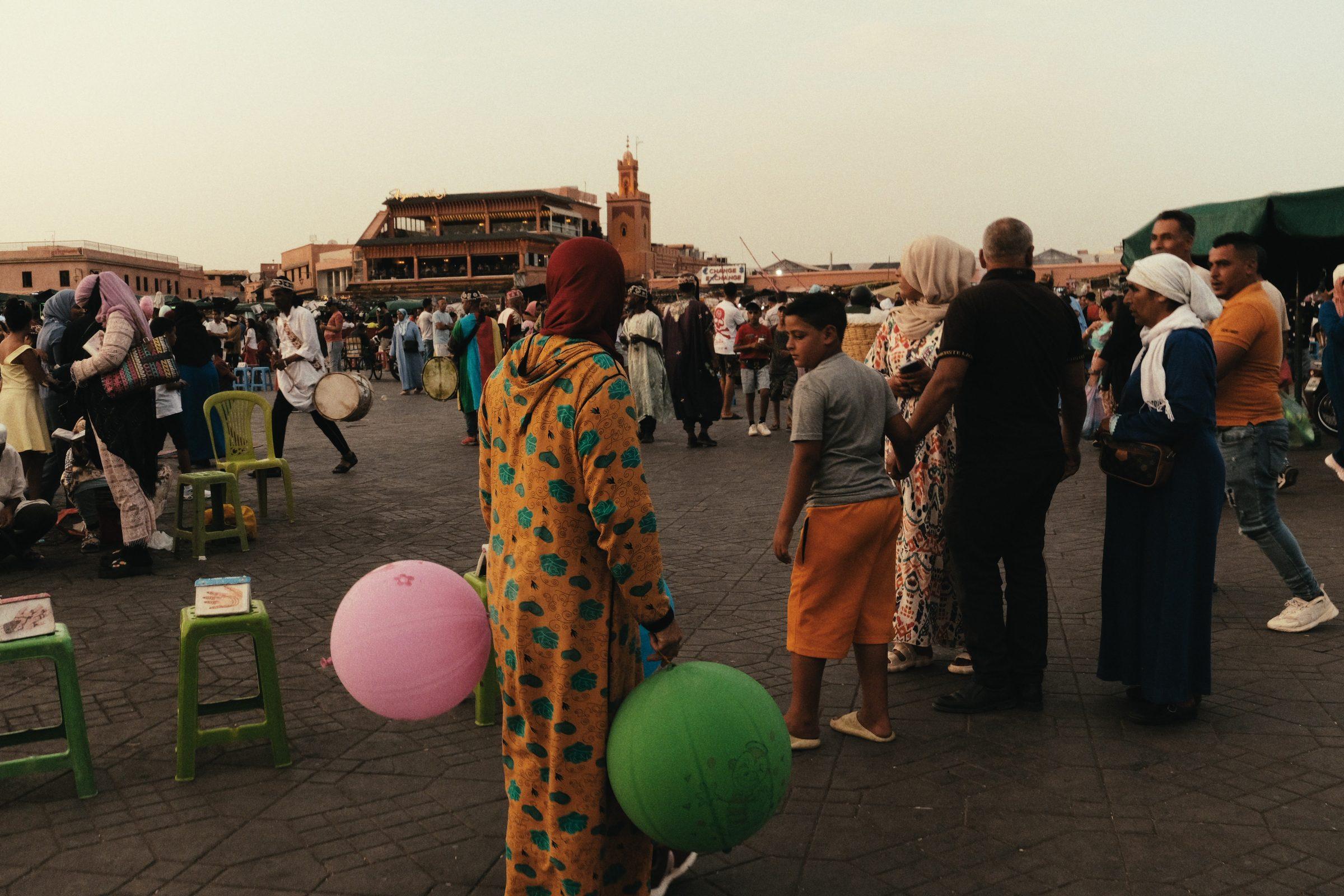 A woman in a patterned dress holding pink and green balloons in the square, a crowd and a minaret behind her