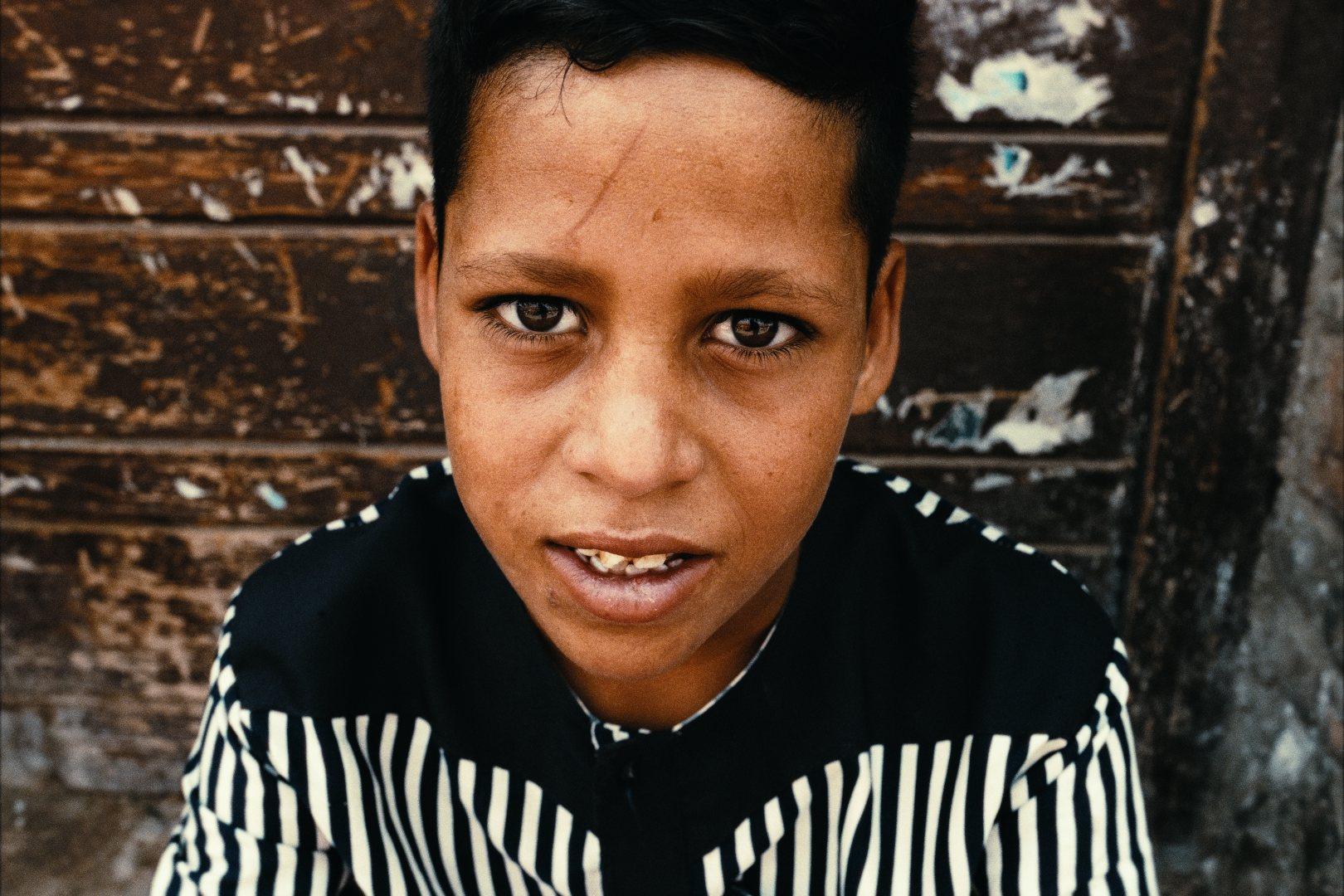 A boy in a black and white striped shirt looking into the camera, in front of a scratched wooden door