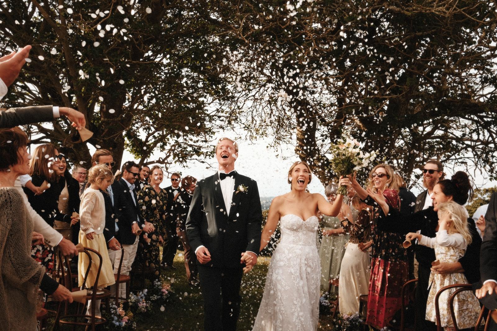 Newlyweds walking through confetti under a tree, surrounded by guests