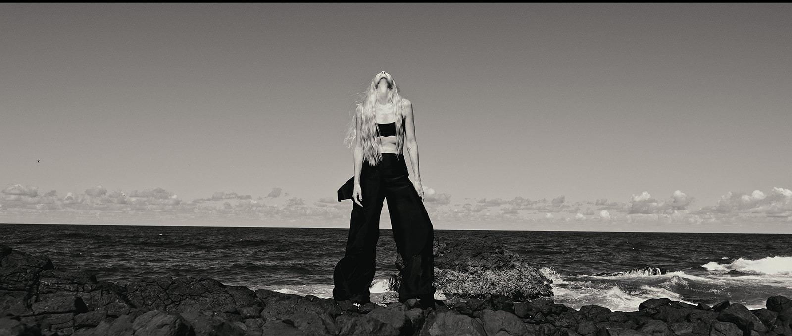 A woman in black standing on the rocks, head tilted back, wide ocean horizon behind her, black and white