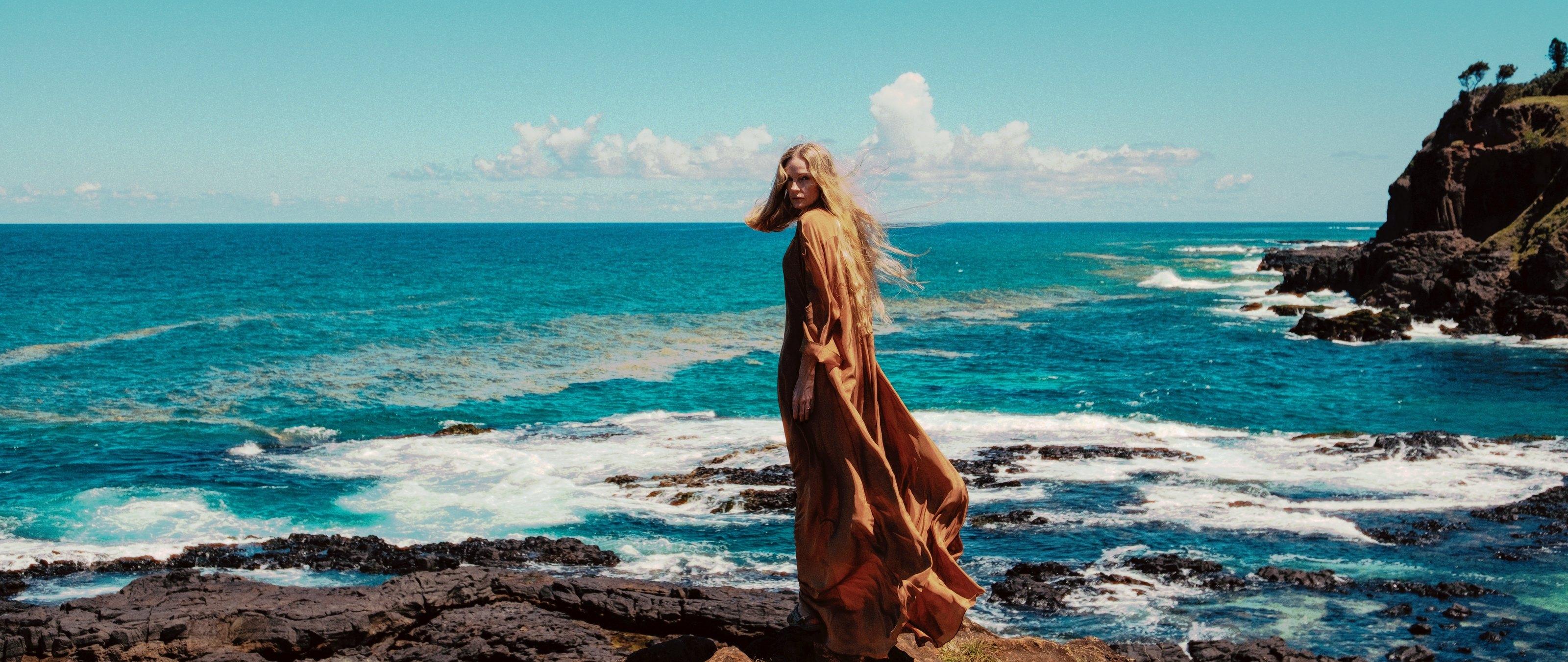 Una donna con un abito color rame che si volta a guardare una costa rocciosa