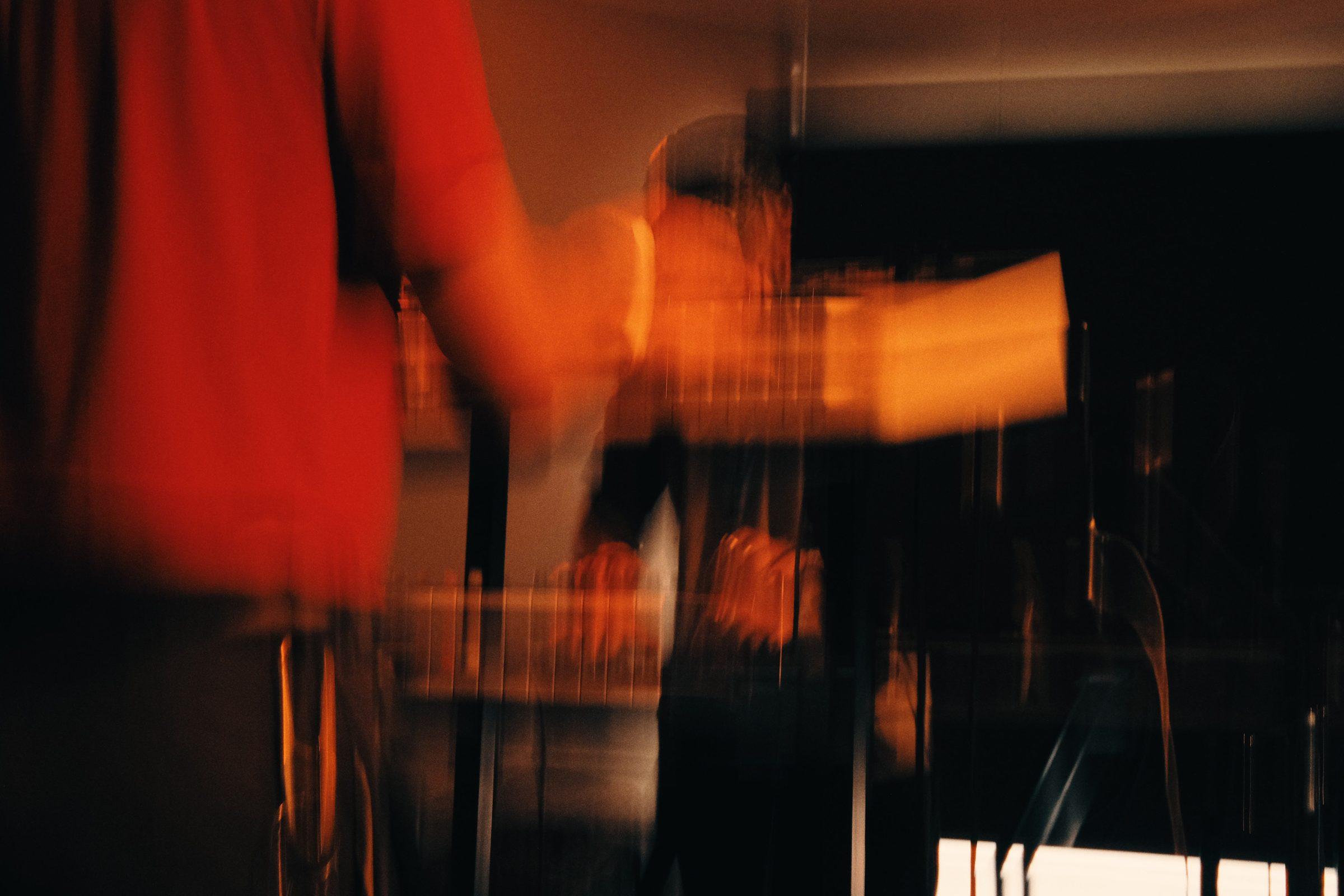 A blurred long exposure of a musician in a red top at a keyboard, in warm stage light