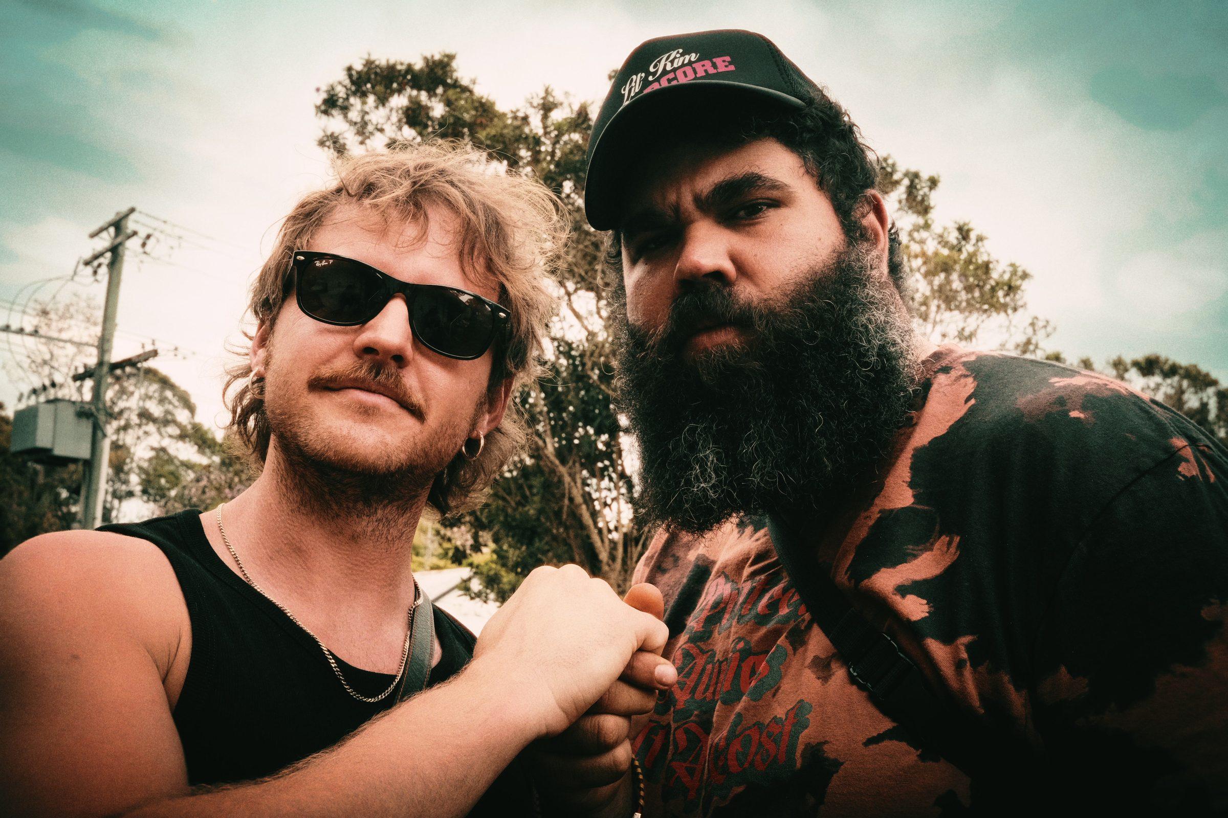 Two men in sunglasses close to the camera, one bearded and in a cap