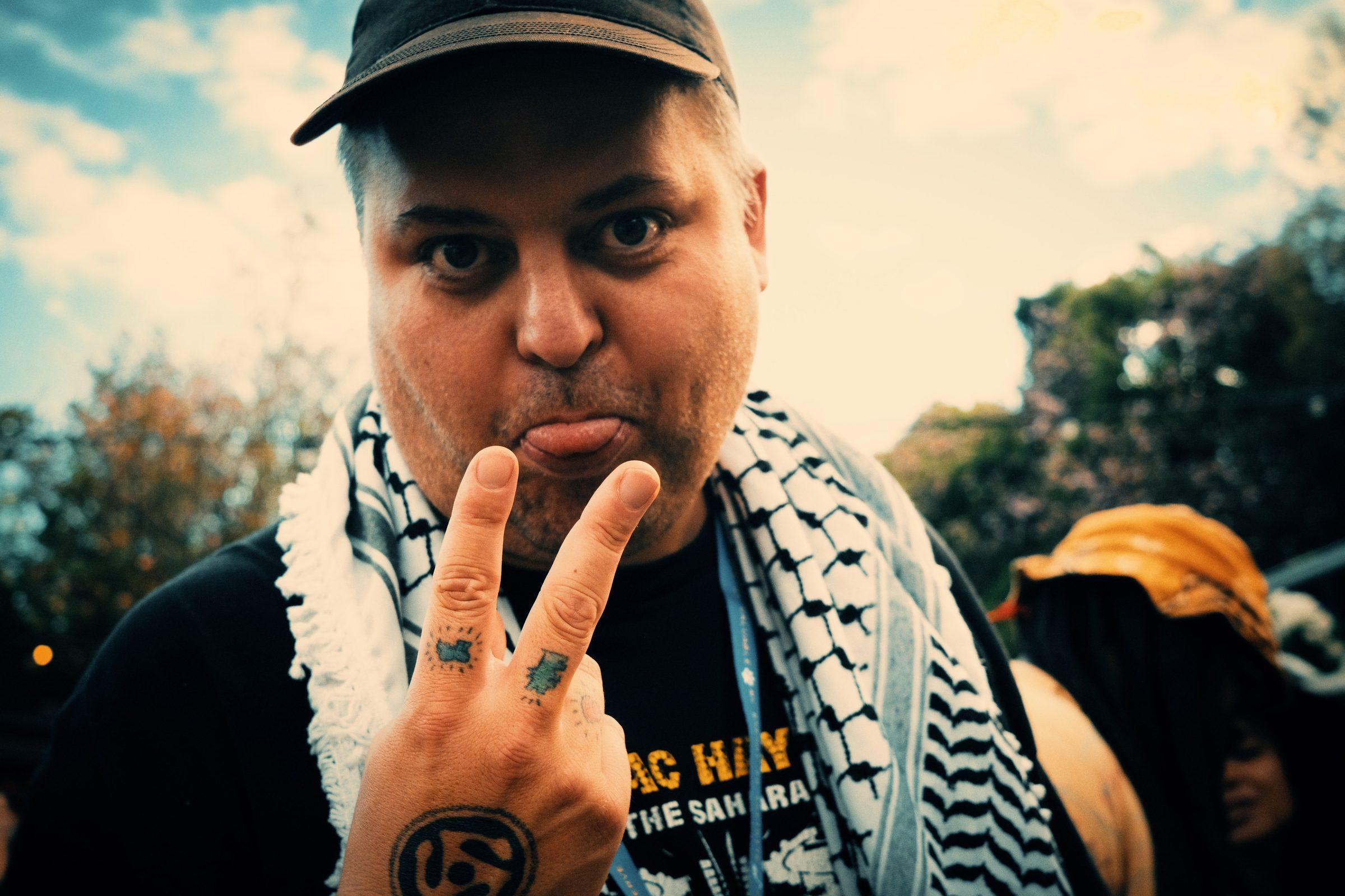 A man in a keffiyeh and cap making a peace sign toward the camera