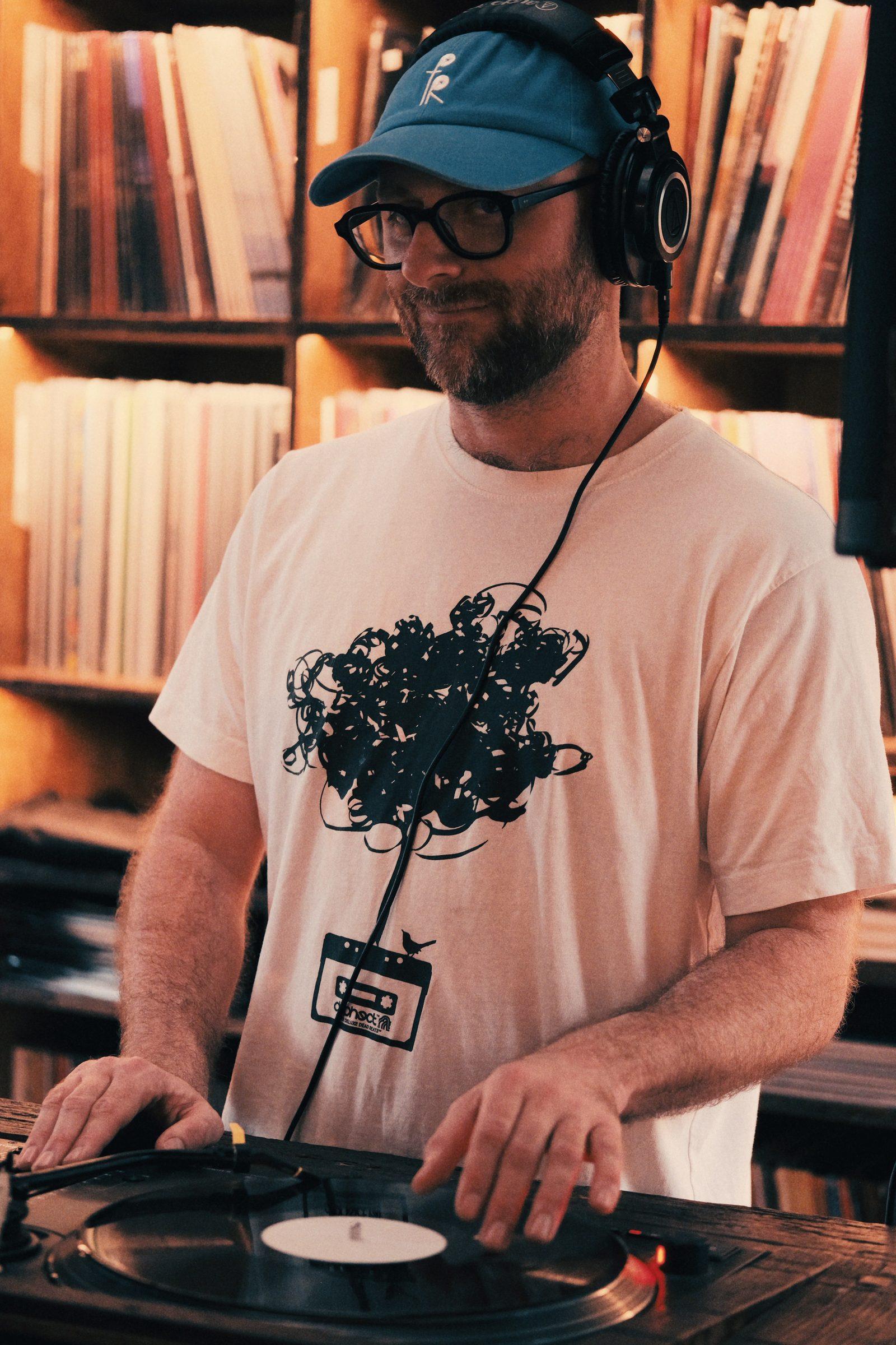 A DJ in a blue cap and headphones at the decks, records on the shelves behind him
