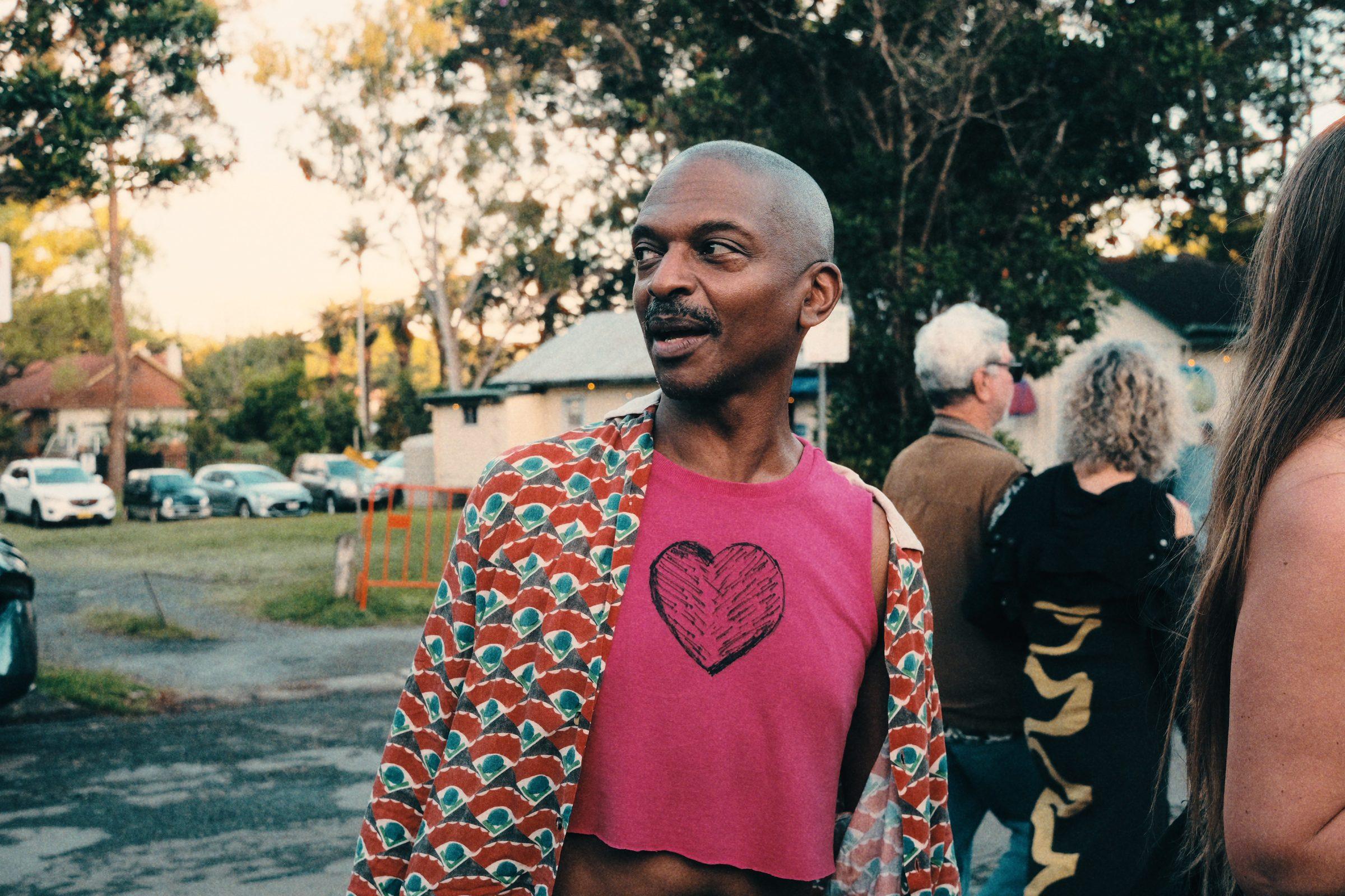 A man in a pink heart-print top looking off to the side, a street behind him