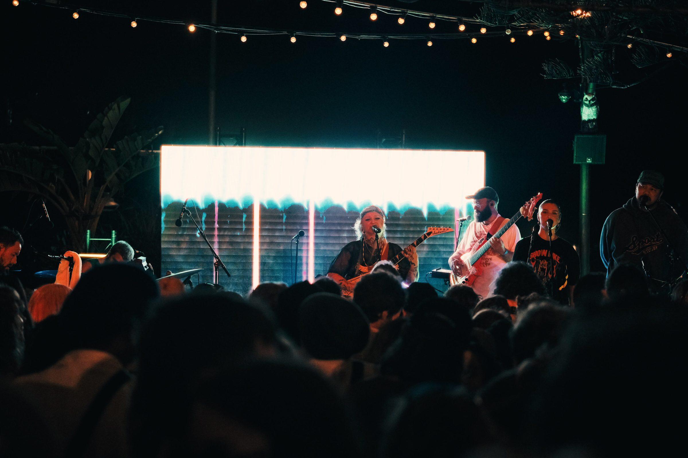 A band on a lit stage in front of a screen, a crowd in the foreground at night