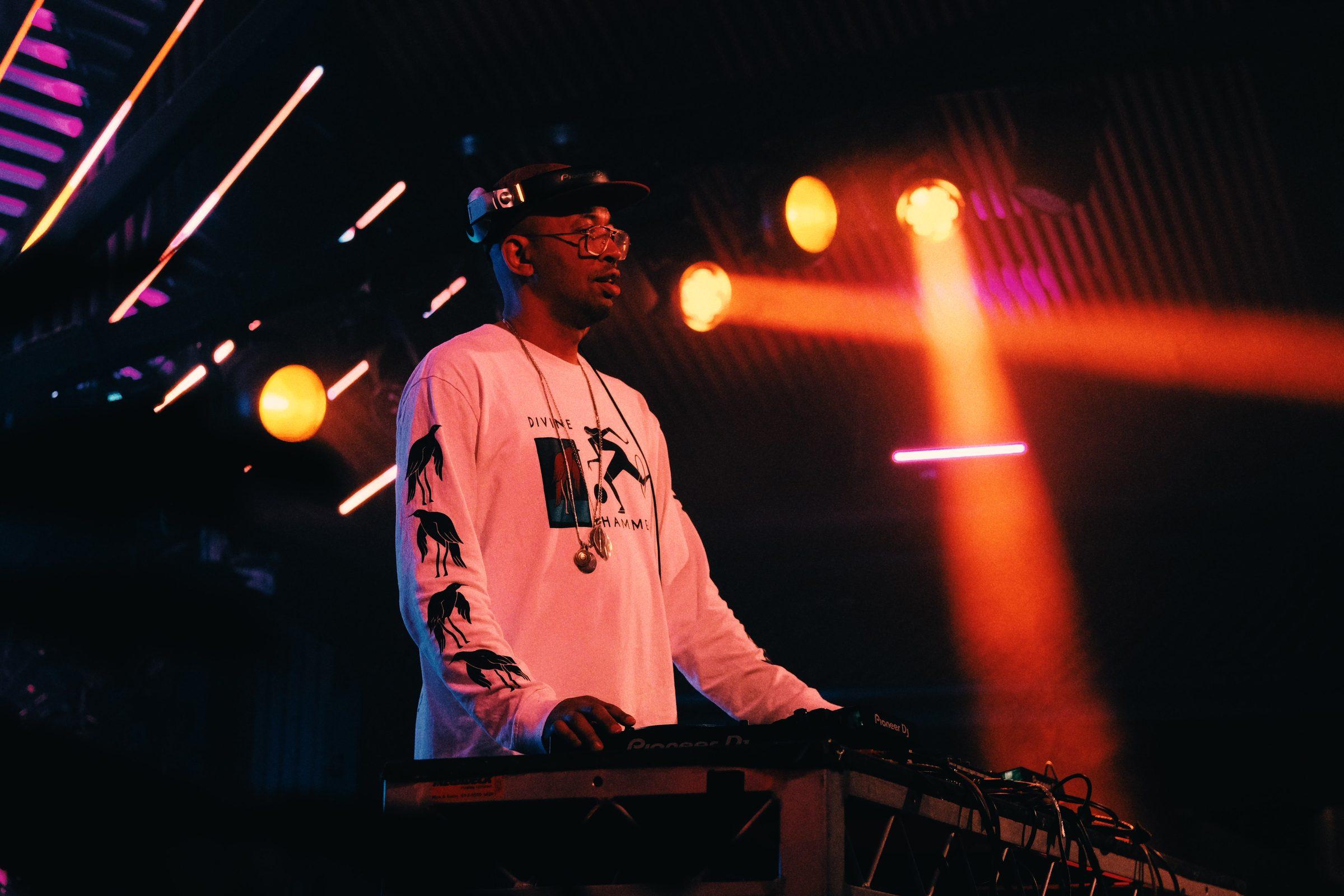 A DJ in a white sweatshirt at a mixing desk, in orange and magenta stage light