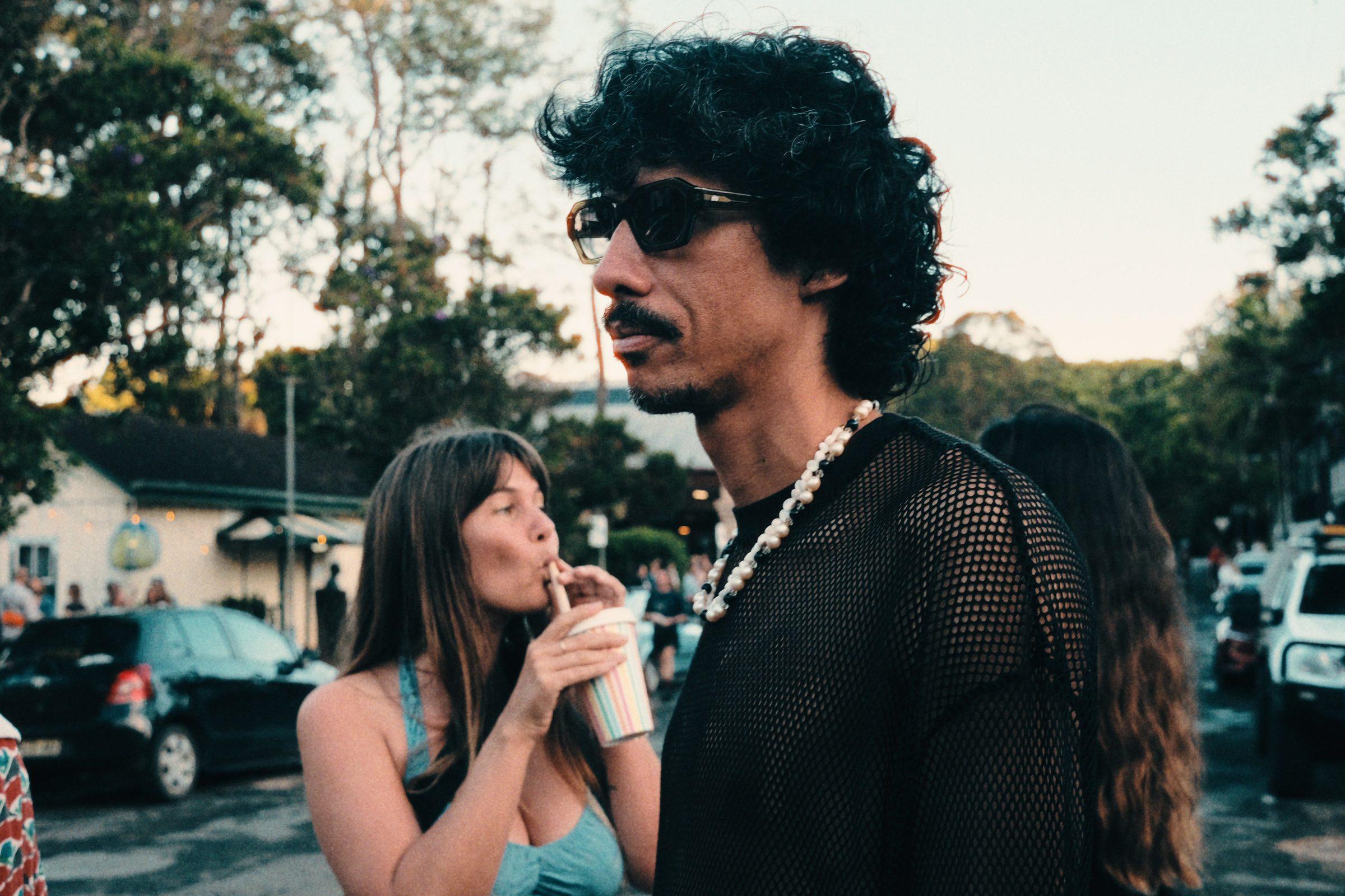 A woman with a drink and a man in sunglasses with dark curly hair, talking on a street
