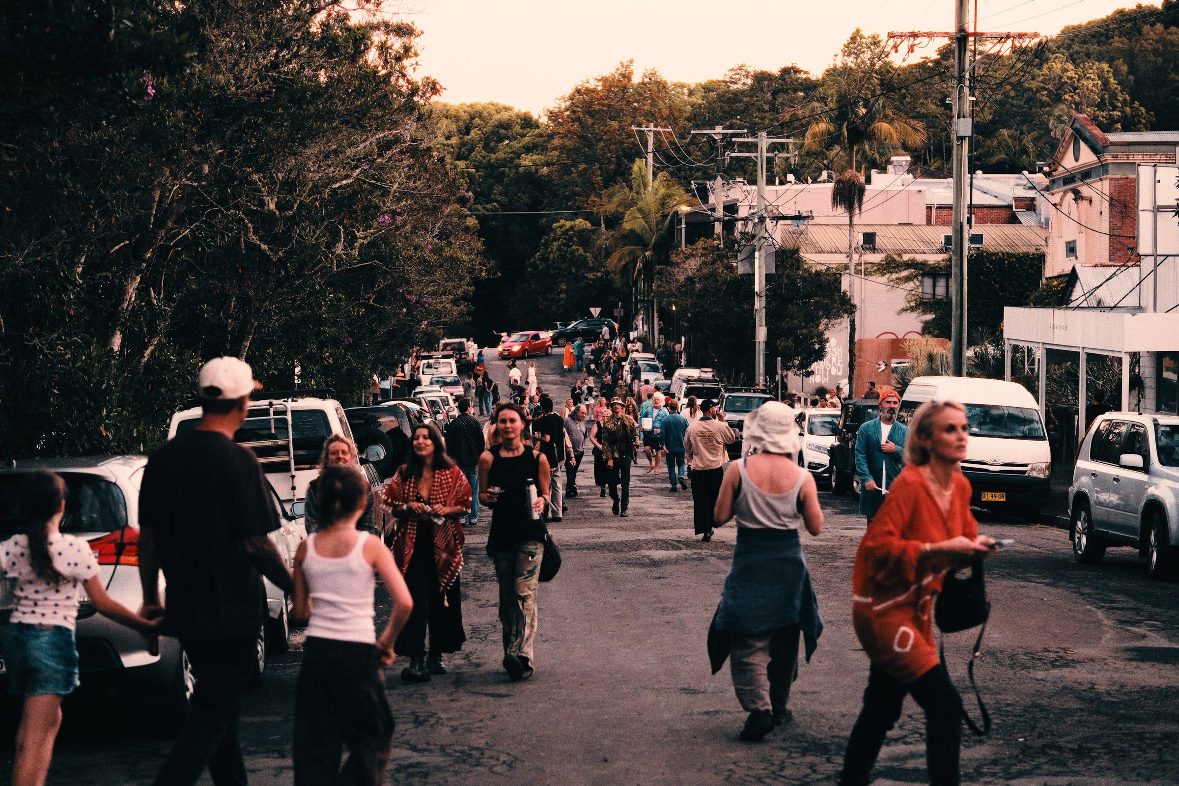 A street full of people walking as the light fades, cars parked on both sides